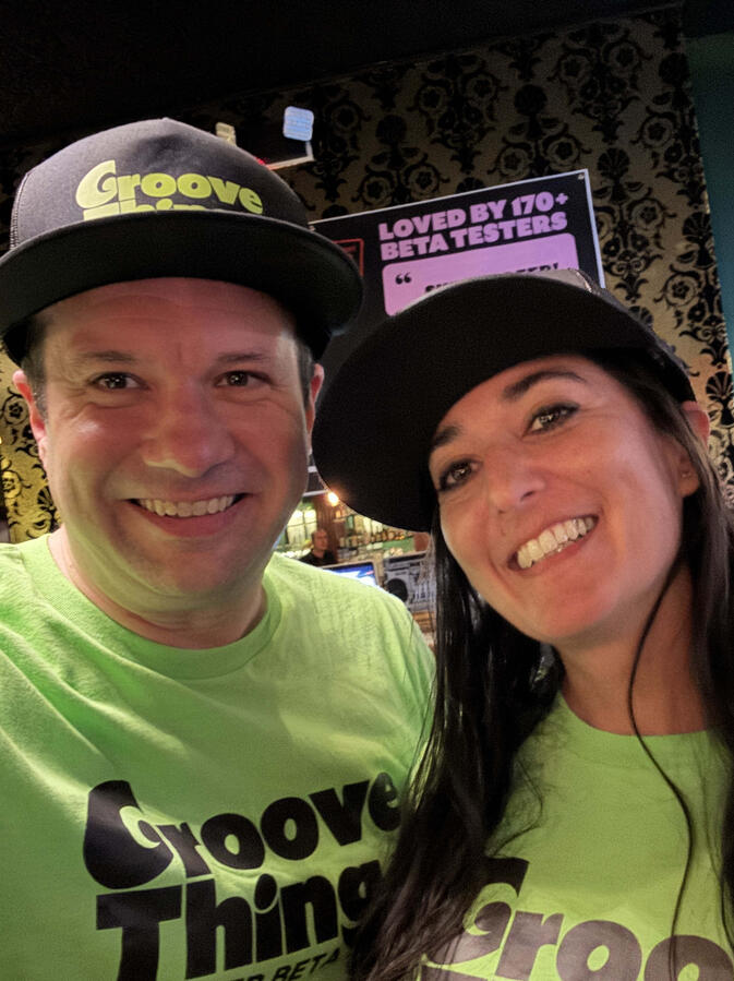 Photo of Michael and Michelle smiling together while wearing "Groove Thing" hats and shirts. Michael and Michelle selfie with red squiggly art in the background.